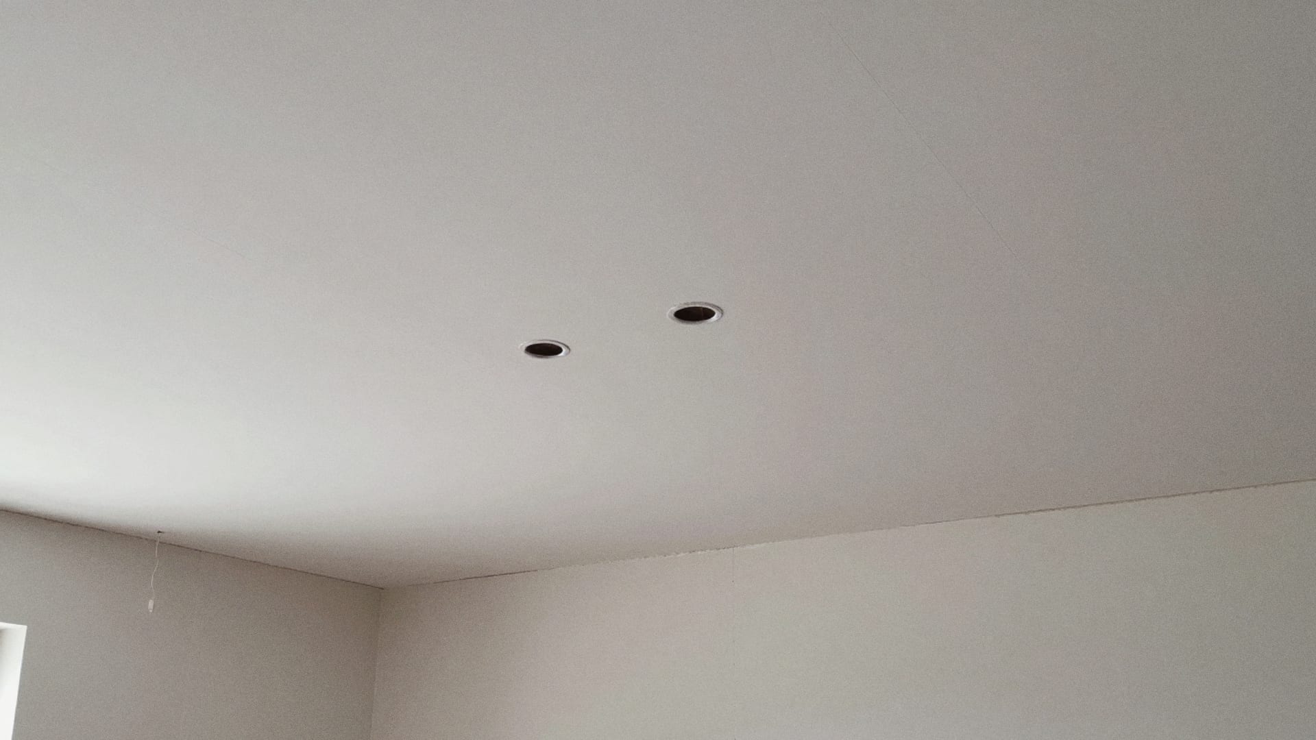 Nieuw wit plafond van gipsplaten met twee ronde gaten voor inbouwspots, gezien vanuit een hoek van de kamer