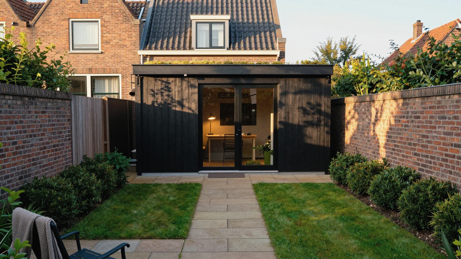Zwart houten tuinhuis met glazen deuren achter in een tuin met gazon en een tegelpad