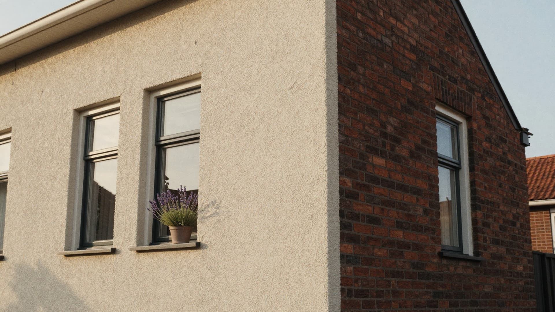 Gevel met beige sierpleister en grijze kozijnen naast een rode bakstenen gevel