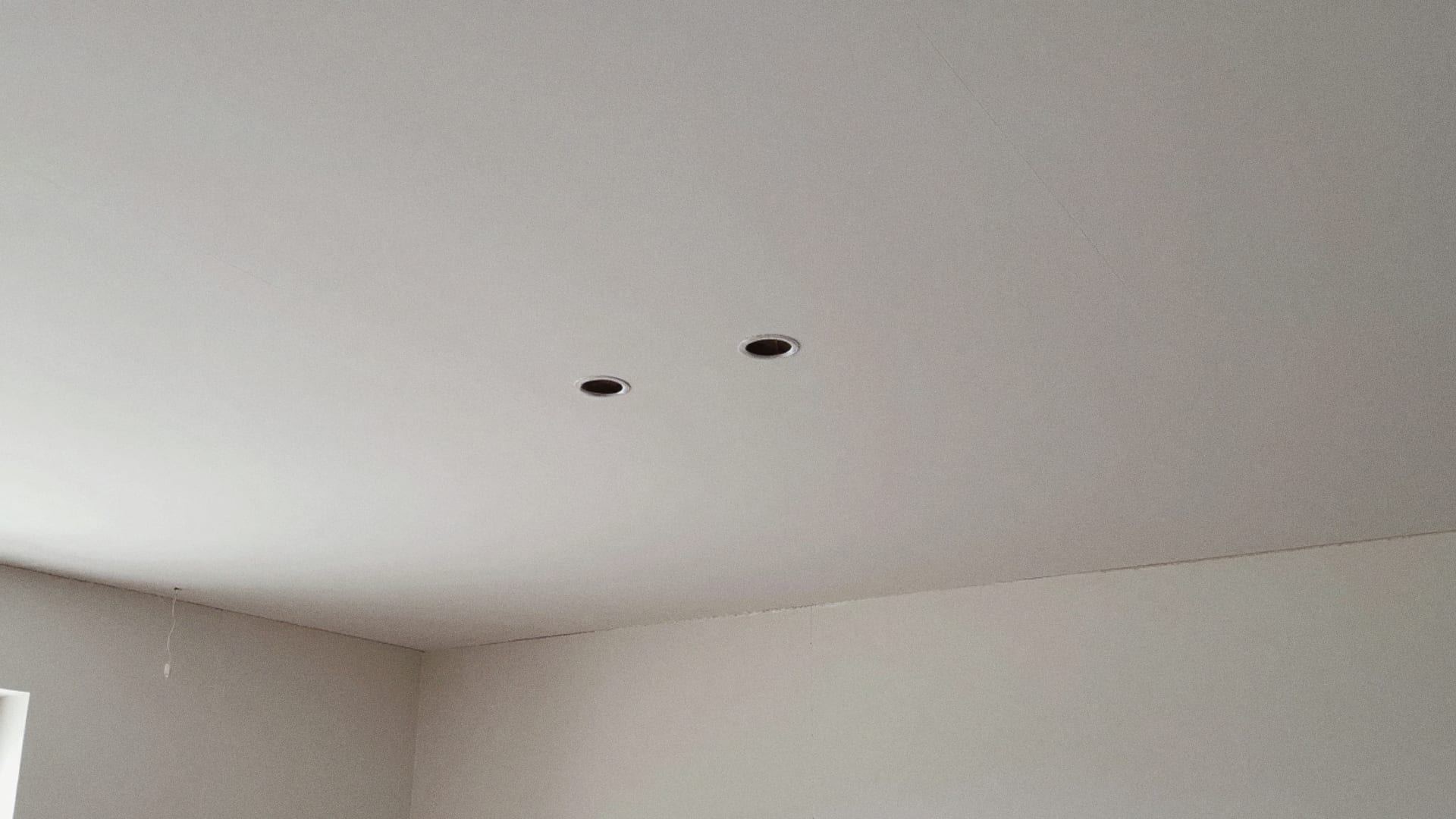 Nieuw wit plafond van gipsplaten met twee ronde gaten voor inbouwspots, gezien vanuit een hoek van de kamer