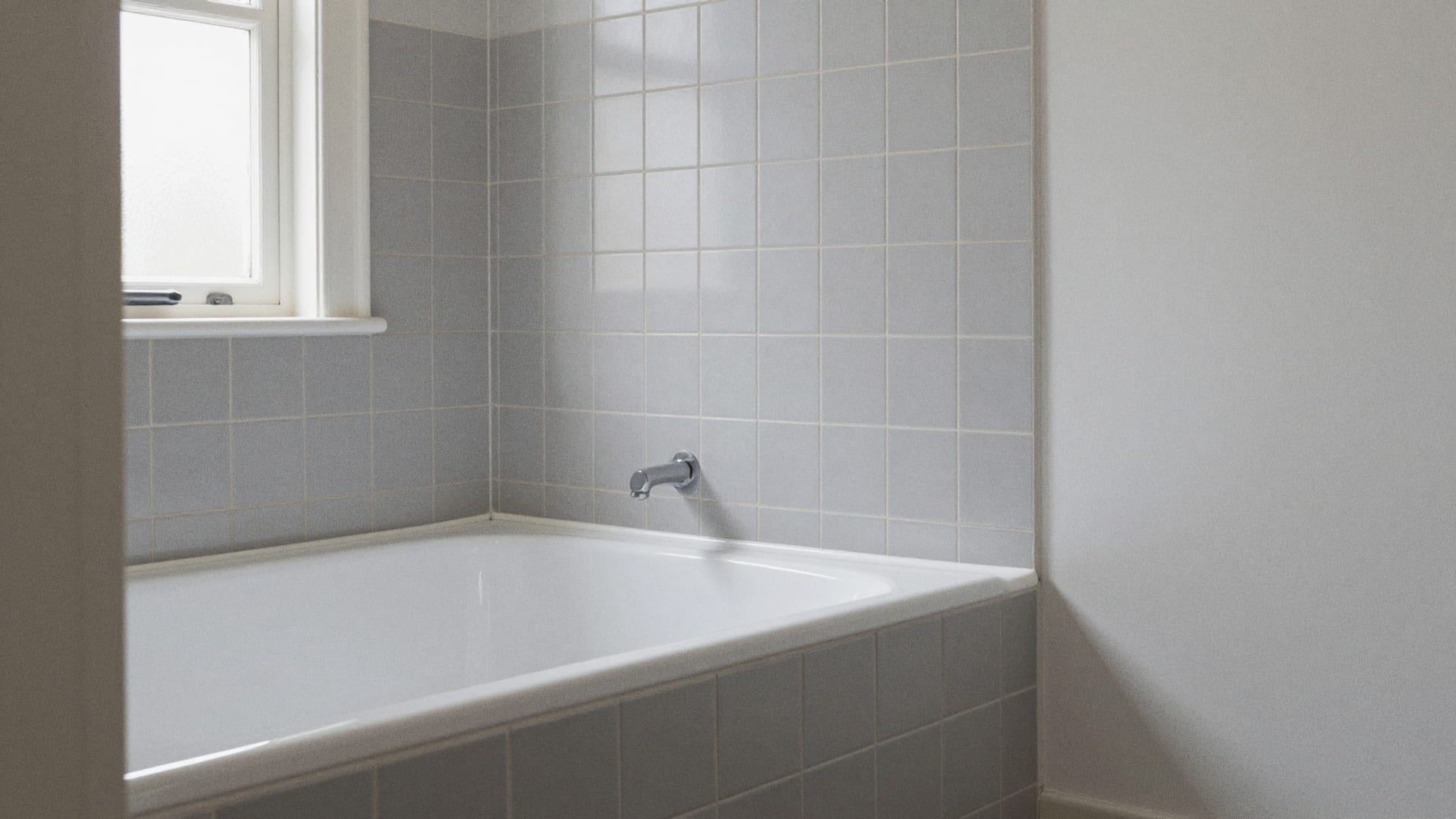 Wit ligbad met een strakke nieuwe witte kitnaad langs een lichtgrijze tegelwand in daglicht